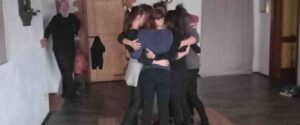 Un gruppo di persone disposte in cerchio che si abbracciano durante una sessione di meditazione in una sala interna.